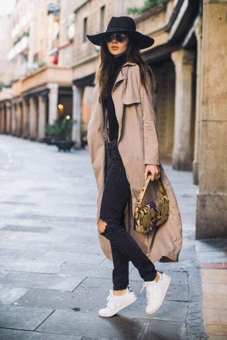 Chica usando sombrero negro, tenis blancos, jeans y blusa negra, junto a una gabardina camel