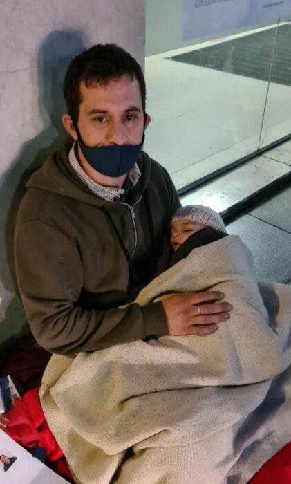 Hombre abrazando a su bebé mientras pide dinero afuera de un banco 