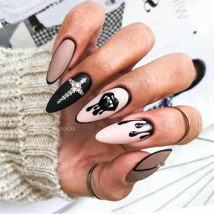Uñas largas stiletto color rosa, negro y nude, con diseño de cruz de pedrería, labios góticos con colmillos de vampiro
