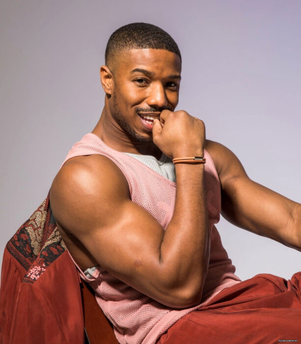 Actor Michael B. Jordan sentado, con una camiseta blanca y rosa de tela de red, pantalón y saco rojo, mostrando sus brazos musculosos, sonriendo con la mano en la boca