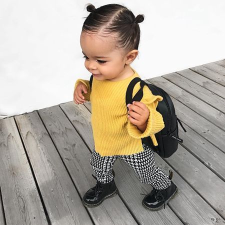 Niña pequeña de cabello negro peinado en chonguitos con suéter amarillo, pantalón de cuadros y zapatos negros