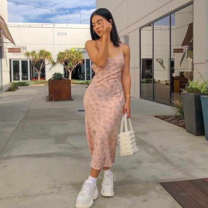 chica con cabello oscuro, vestido slip rosa con flores, bolsa de cuentas blanca y tenis deportivos chunky blancos