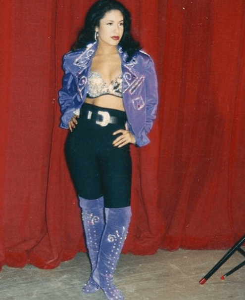 Selena Quintanilla usando leggins negros con botas moradas, bustier de pedrería y chaqueta morada 