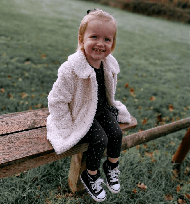niña de cabello rubio con ojos azules, usando una sudadera negra con lunares blancos, pantalón negro con lunares blancos, tenis converse negros y abrigo teddy color beige