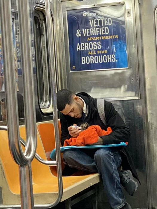 Hombre sentado en el metro con saco negro, camisa blanca, corbata negra y jeans, alimenta a un gatito café con rayas negras, mientras lo arropa con una manta naranja y le da de comer con un biberón