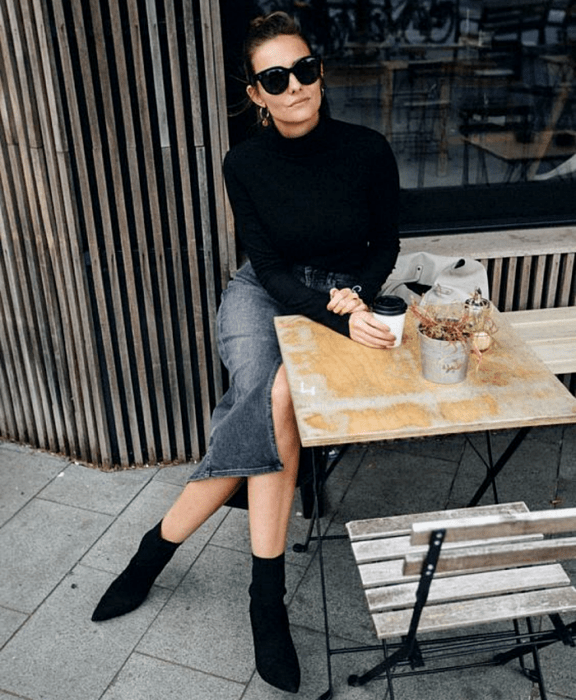 mujer con cabello castaño usando lentes de sol, suéter negro de cuello alto, falda midi de mezclilla y botas tipo sock boot negras