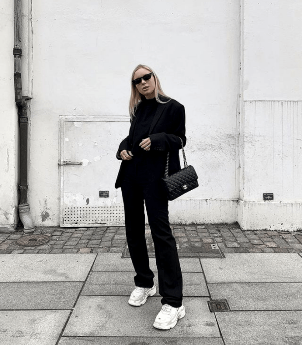 chica rubia con lentes de sol, camiseta negra, saco negro, pantalones de vestir negros, tenis deportivos blancos y bolso de mano negro