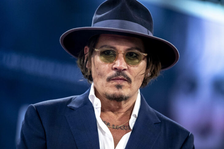 Actor Johnny Deep usando camisa blanca, saco y sombrero con unos lentes de sol