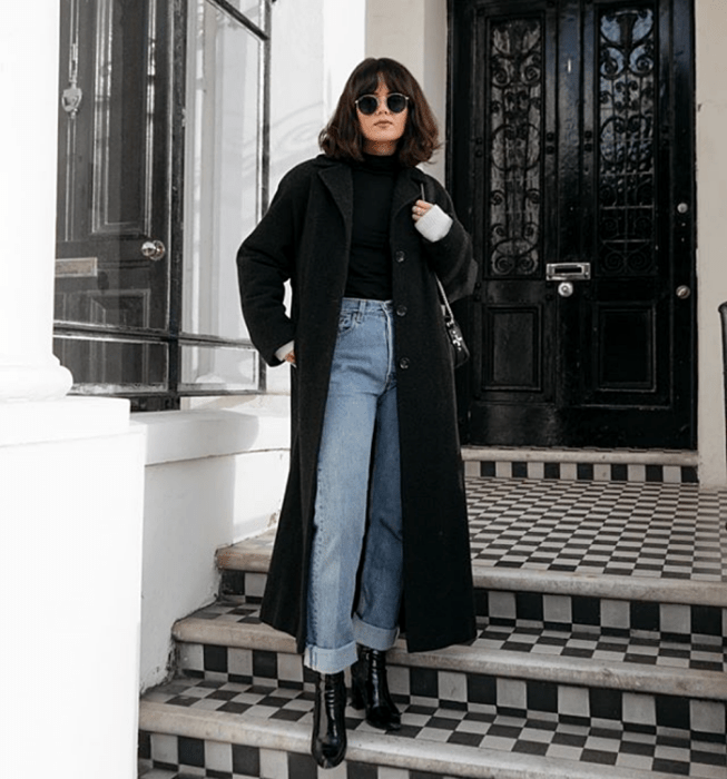 mujer de cabello castaño oscuro usando lentes de sol, blusa negra de manga larga, abrigo largo negro, jeans rectos a la cintura, botas negras de tacón y bolso negro