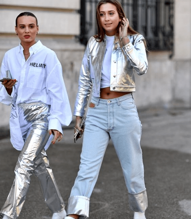 chicas de cabello castaño, una usa una camiseta blanca con estampado, pantalones plateados metálicos y botas blancas, la otra lleva una camiseta blanca crop top, chaqueta plateada metálica, jeans a la cadera con detalles metálicos y botas blancas