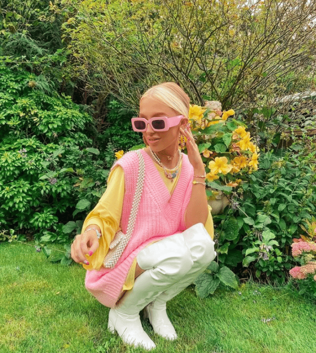 joven rubia con gafas de sol rosas, suéter rosa con mangas amarillas, botas largas blancas de cuero y bolso rosa