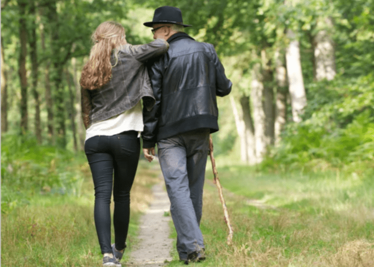 una hija camina con su papá por un camino lleno de vegetación, el hombre lleva sombrero y bastón, ella viste casual