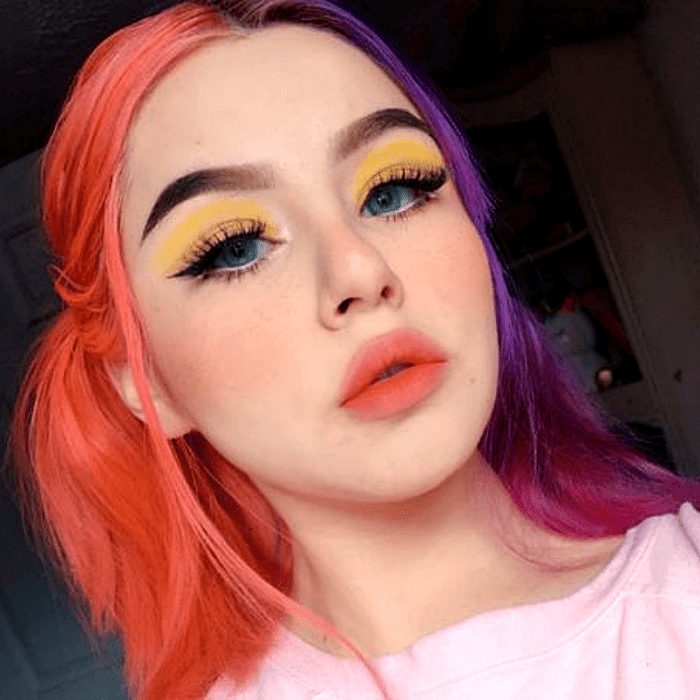 chica de ojos azules con cabello teñido en color naranja y morado neón, maquillaje atrevido, sombras amarillas y delineador negro