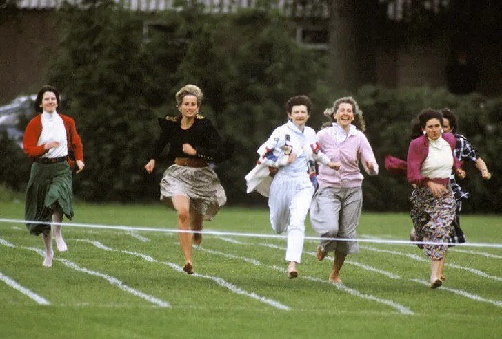Lady Di usando una falda de color café con un saco negro y descalza corriendo junto a otras madres en un evento escolar