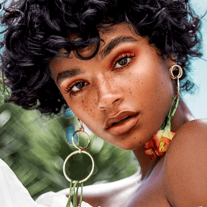chica morena de cabello chino, con pecas, usando maquillaje con sombra roja, rimel negro y aretes de flores