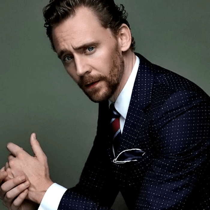 tom hiddleston con traje azul, camisa blanca de vestir y corbata roja