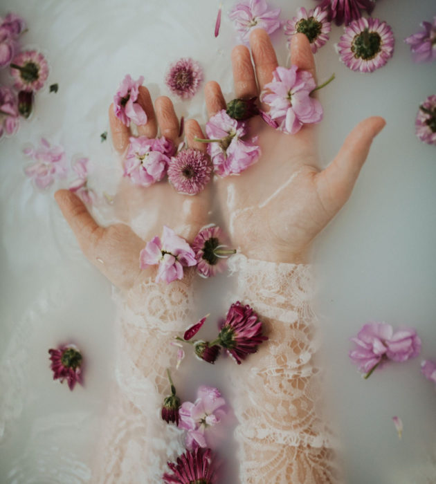 Milk bath; fotografía de manos de mujer en el agua con leche y flores moradas