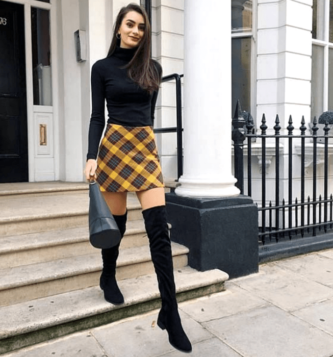 Chica de cabello castaño largo luciendo un top negro de cuello alto y manga larga, minifalda de cuadros escocesa en amarillo y azul, botas largas negras de tacón bajo y bolso de mano de cuero