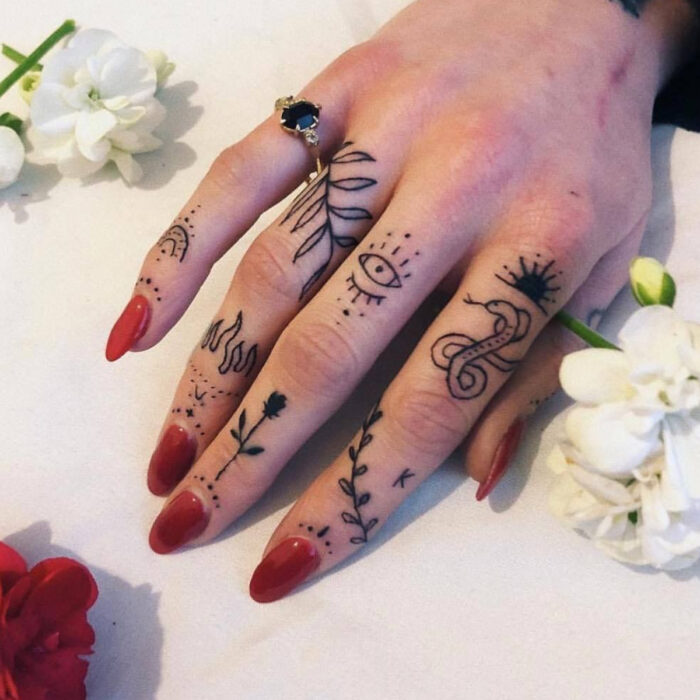 Manos elegantes con manicura, uñas largas en forma de almendra con esmalte rojo, dedos tatuados con ojos, llamas, flores, serpiente, hojas y sol, anillo con piedra negra, flor blanca y roja
