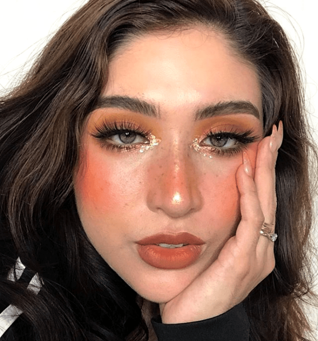 chica de cabello castaño usando sombra naranja, pestañas postizas, rubor intenso, labial naranja oscuro y brillo dorado en el lagrimal