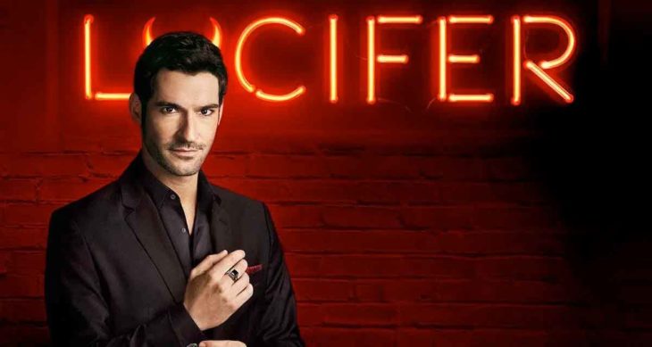Tom Ellis, actor, ajustando el puño de su camisa negra, posando de perfil y vistiendo un traje oscuro.